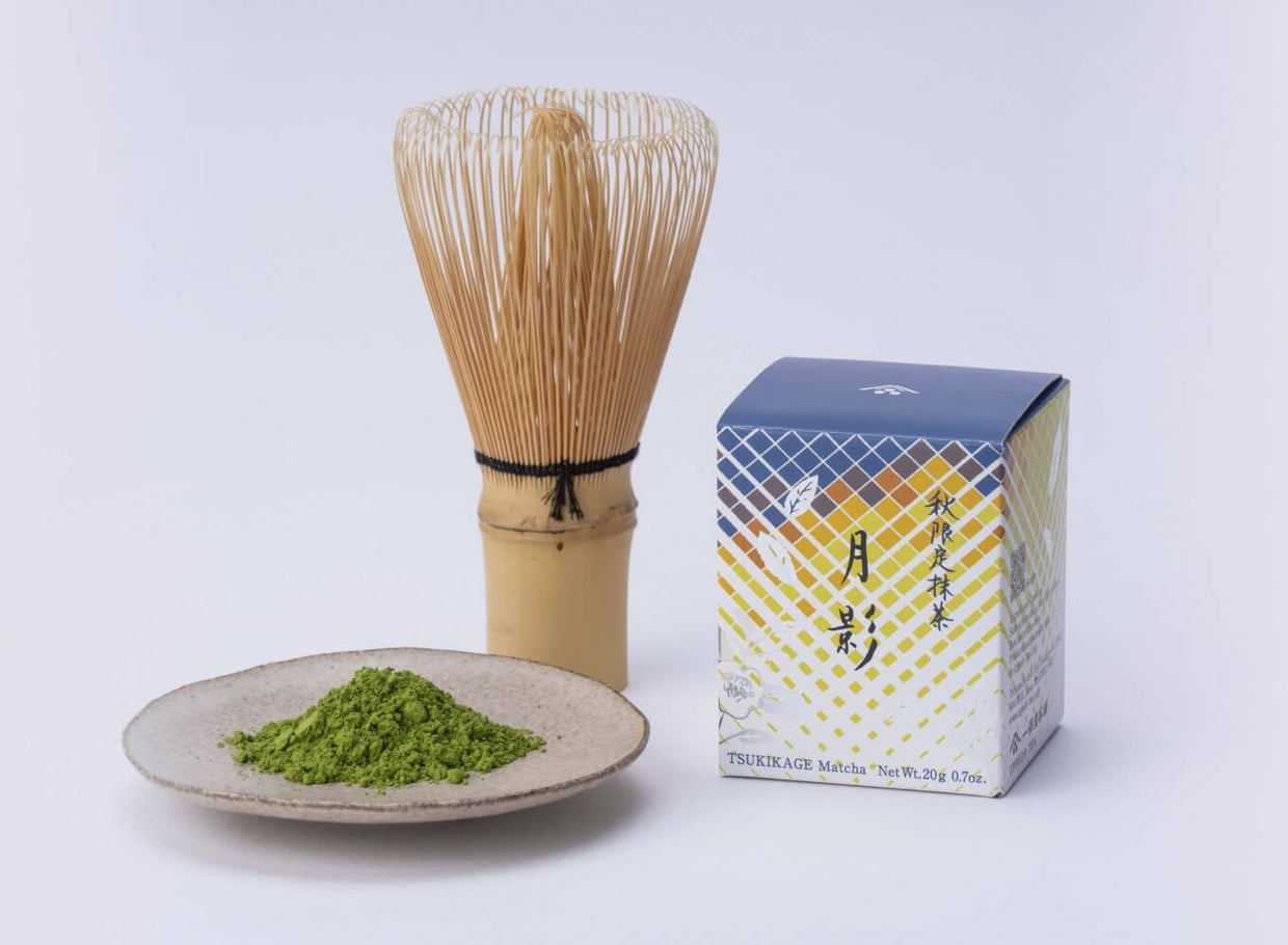 A product photo of Tsukikage fall matcha, showing the box, a bamboo whisk, and a pile of vibrant green matcha powder on a ceramic plate against a white background.