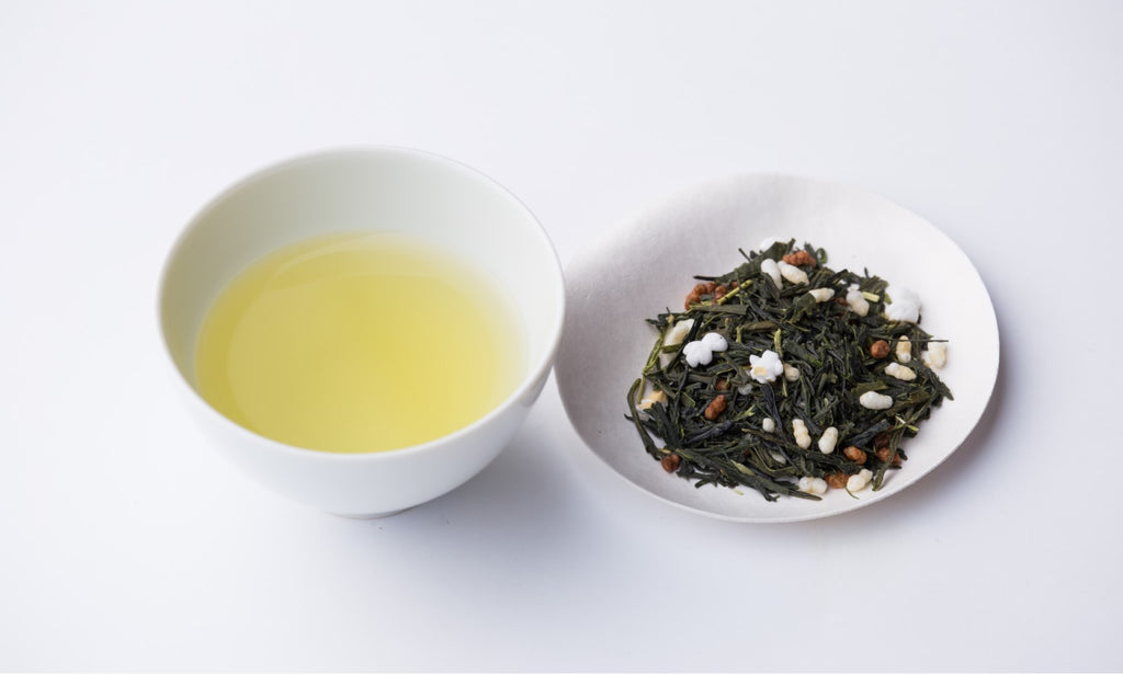 White teacup of light Genmaicha green tea with roasted rice beside small plate of dried bancha tea leaves with roasted rice
