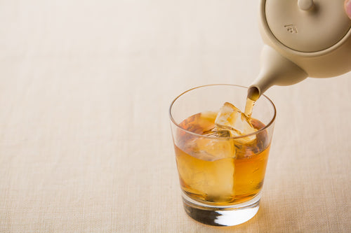 Pouring hot amber tea from ivory banko-yaki clay kyusu teapot into glass containing several ice cubes and roasted Hojicha tea