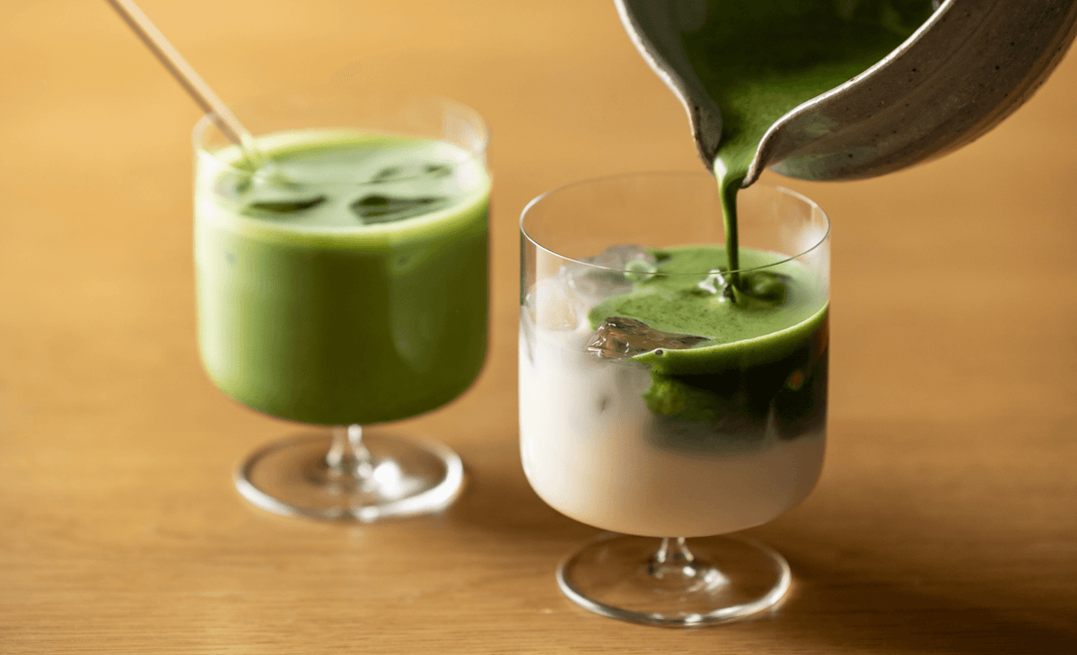 Pouring vibrant green matcha float from spouted bowl over milk and ice in elegant stemmed glassware beside iced matcha latte
