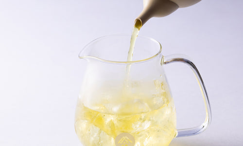 Green tea being poured from elegant spout of ivory teapot into small rounded modern glass pitcher with white Ippodo Tea logo