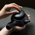 Hands opening a black Ippodo Tea kyusu. Round Japanese teapot with left side handle and lid. Green tea preparation.
