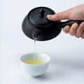 Hand pouring green tea from a black Tokoname-yaki kyusu teapot with side handle Ippodo Tea, filling a white teacup.