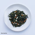 Sample of Ippodo Genmaicha dried bancha green tea leaves and stems mixed with roasted rice on white plate, on white table