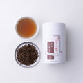 Can of Ippodo Tea Hojicha beside porcelain teacup of amber color roasted green tea and plate of roasted loose leaf and stems - thumbnail 4