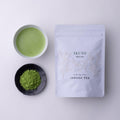 Teacup of green prepared matcha tea beside black plate of loose tea powder and white resealable bag of Ippodo Ikuyo matcha 