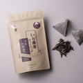 Tan and burgundy tea package with loose tea leaves and two pyramid-shaped tea bags on a light pink background