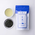 Teacup of brewed sencha green tea alongside silver plate of loose rolled tea leaves and blue packaging for Kumpu Ippodo Tea