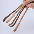 Hand holding a set of five wooden tea ladles with various finishes and colors against a light gray background
