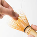 Holding Korean artisan-made Chasen 80-tip bamboo matcha whisk with fingers and rinsing with water