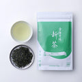 Teacup of light yellow brewed shincha tea beside silver plate of loose rolled tea leaves and green package of New Harvest Tea