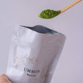 Scooping spoonful of bright green matcha powder out of large white reseleable 40g bag of Ummon by Ippodo Tea