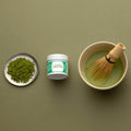 Green matcha tea powder on silver plate beside tin of Organic Matcha and bowl of prepared matcha with whisk on green table