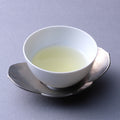 Small white porcelain cup of light color Japanese green tea on silver dual petal leaf shaped saucer on grey table - thumbnail 3