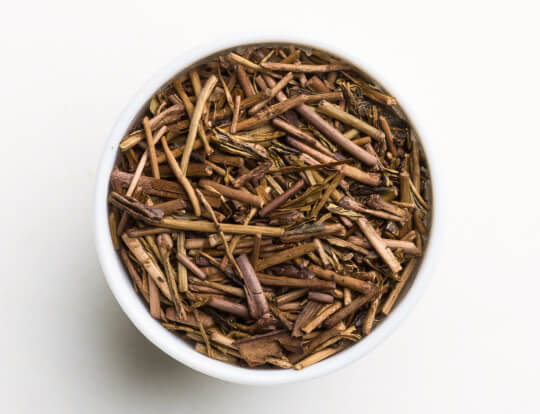 Loose dried brown Ippodo Tea Co. Stems (Kuki) Hojicha Japanese roasted green tea stems in white bowl on white background