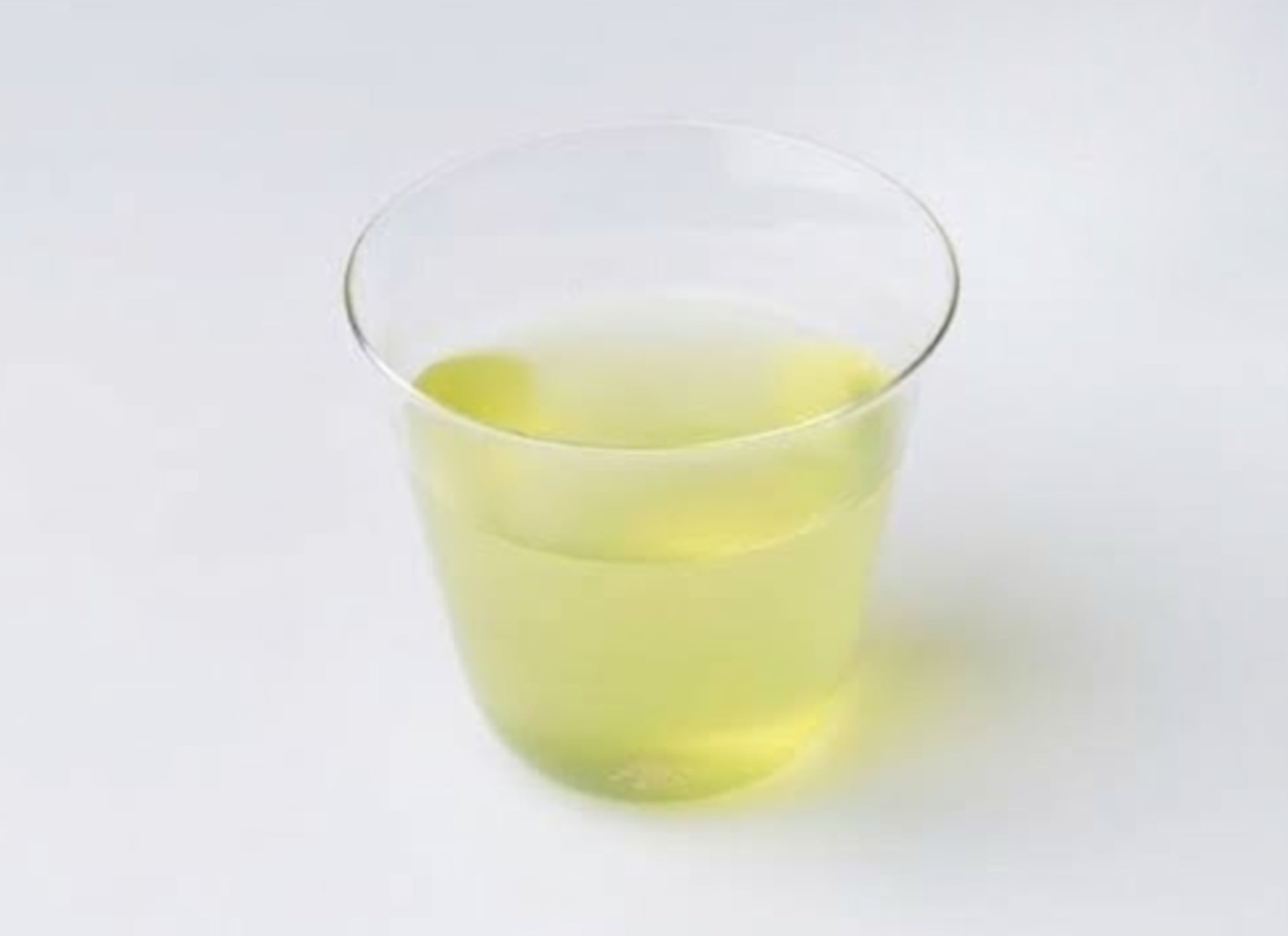 A tea bag hovering over a clear glass teapot/pitcher with a metal handle, photographed on a white background. The genmaicha tea bag appears to contain loose tea leaves and what look like small dried fruits or flowers. - Genmaicha Teabags - Cold Brew