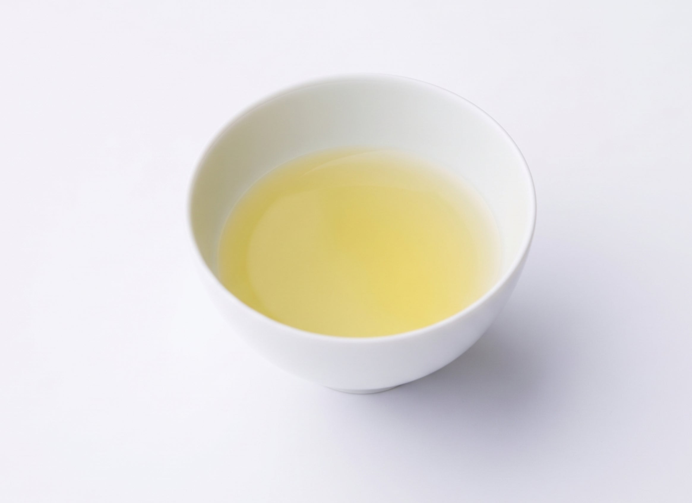 A hand pouring pale yellow-green tea from a glass teapot into a small glass cup. The teapot has a metal lid and handle, and contains what appears to be a pyramid tea bag. There's a logo visible on the teapot that reads "IPPODO TEA". - Genmaicha Teabags - Hot Brew