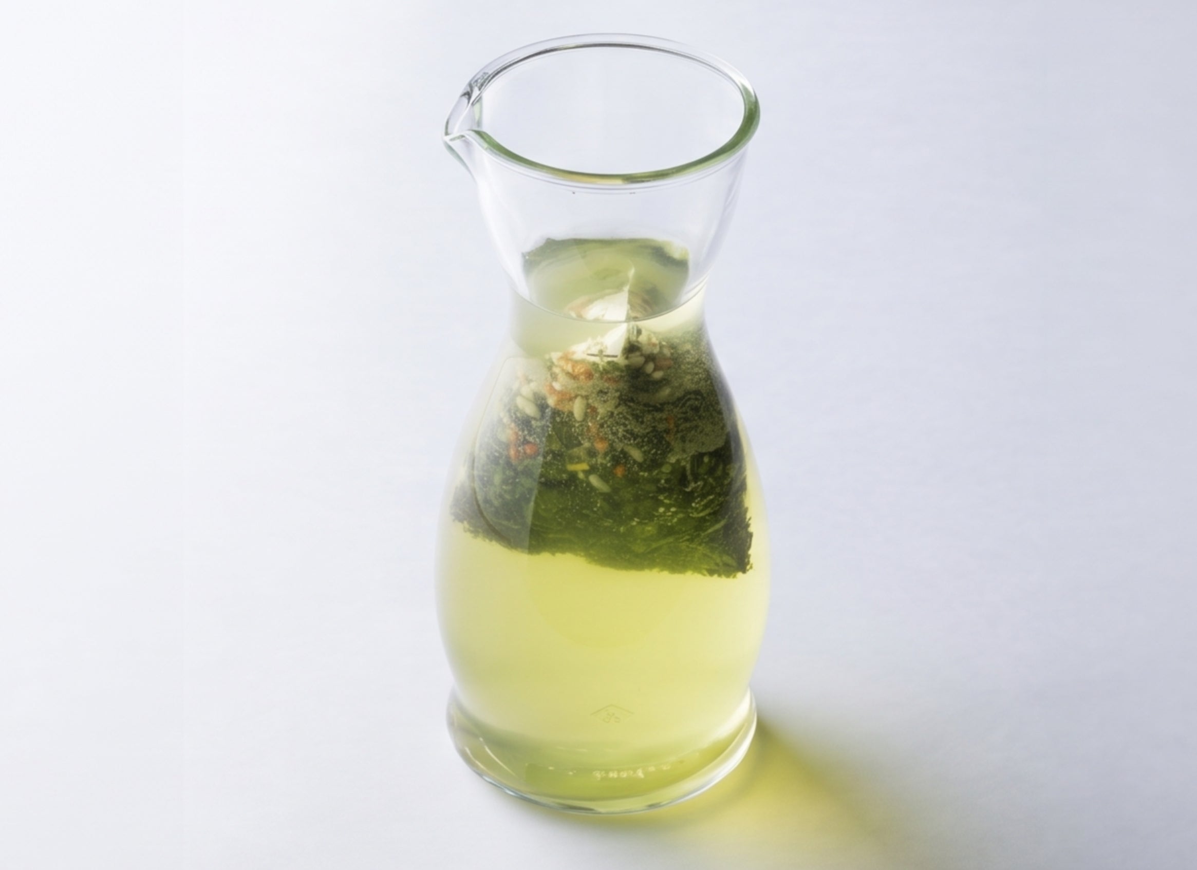 A hand pouring pale yellow-green tea from a glass teapot into a small glass cup. The teapot has a metal lid and handle, and contains what appears to be a pyramid tea bag. There's a logo visible on the teapot that reads "IPPODO TEA". - Genmaicha Teabags - Hot Brew