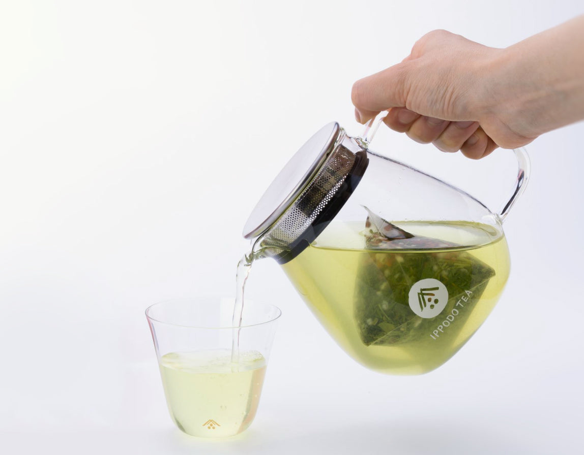 A tea bag hovering over a clear glass teapot/pitcher with a metal handle, photographed on a white background. The genmaicha tea bag appears to contain loose tea leaves and what look like small dried fruits or flowers.