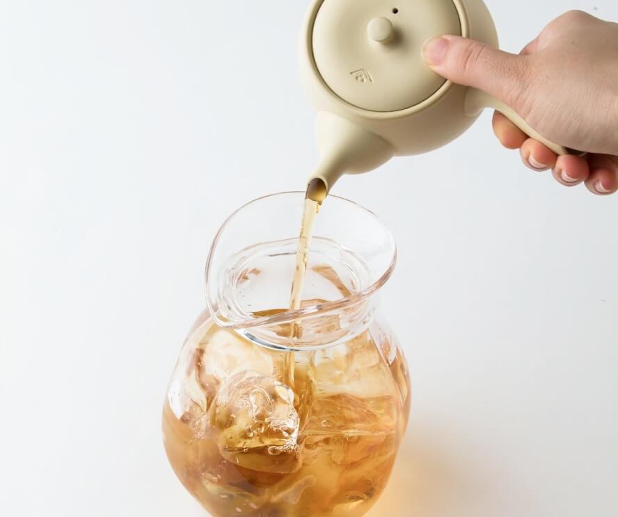 Image of a hand pouring golden-brown tea from a pale ceramic teapot into a glass pitcher filled with ice cubes. The teapot is light beige in color with a simple, rounded design. The clear glass pitcher allows the ice and tea to be visible as it's being poured. The background is white, creating a clean, minimalist composition that emphasizes the action of preparing iced tea. The warm color of the tea contrasts nicely with the cool tones of the ice and glass, creating an appealing visual effect.