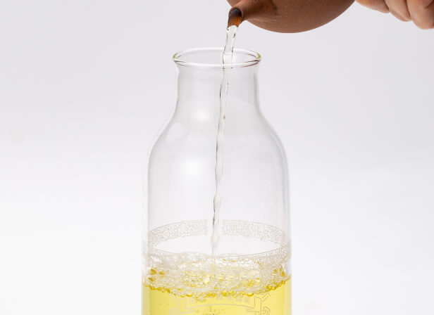 Pouring green tea from above from brown Yakishime clay kyusu teapot into large glass carafe with white Ippodo Tea engravings