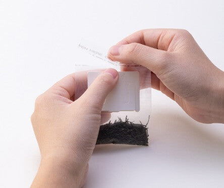 Two hands holding translucent Drip Teabag with bright green leaves inside and removing seal to be able to spread open teabag