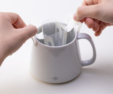 Two hands spreading two panels at top of Drip Teabag across edges of white Ippodo Tea logo mug and clipping teabag to sides