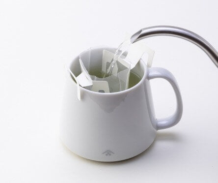 Water being poured from spout of silver teapot into white Ippodo Tea logo mug through Drip Teabag clipped onto top of mug