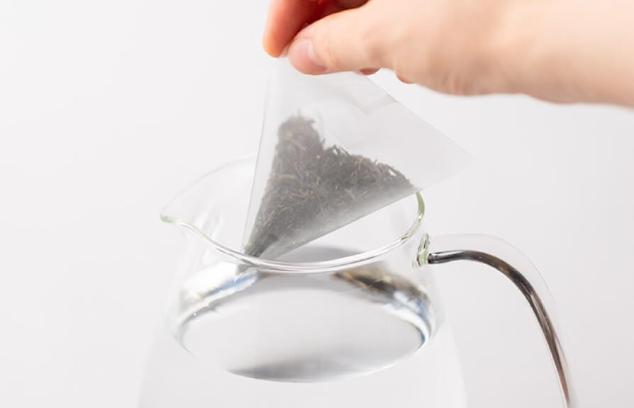 Hand placing large pyramid-shaped teapot-size tea bag into glass pitcher of water.