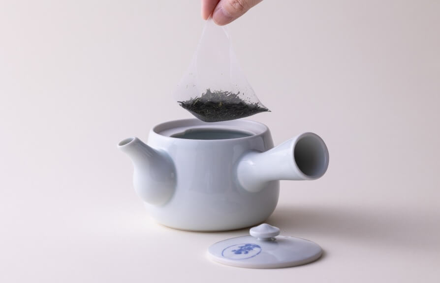 Hand adding large tea-pot size pyramid-shaped tea bag to large white porcelain Kyusu teapot for steeping.