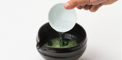 Pouring hot water from white porcelain teacup into black Ippodo Tea Bowl with Spout containing vibrant green sifted matcha